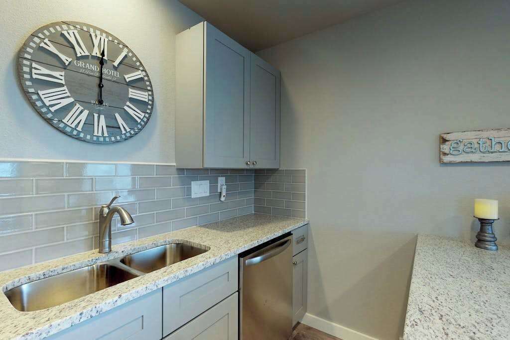 A kitchen with a sink, faucet, and dishwasher, a clock with Roman numerals, and a cabinet above the sink.