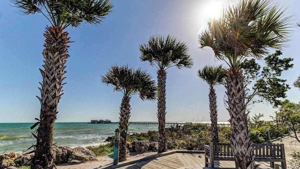 A wooden boardwalk leading to a beach with palm trees and a pier in the background. - Updated bayside condo with heated pool - across the street from the beach (Anna Maria)