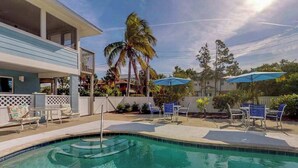 A swimming pool with lounge chairs and umbrellas, surrounded by a building with a balcony and palm trees. - Updated bayside condo with heated pool - across the street from the beach (Anna Maria)
