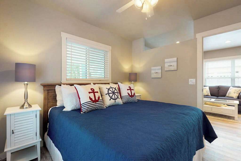 Une chambre à coucher comprenant un lit, une table de chevet, une lampe, une fenêtre avec des stores et donnant sur une autre pièce.