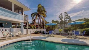 A swimming pool with lounge chairs and umbrellas, a two-story building with a balcony, and palm trees. - Stylish bayside condo with WiFi & pool - close to the beach & restaurants (Anna Maria)