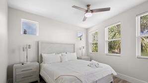 Un dormitorio con cama, mesitas de noche, ventilador de techo y ventanas con vistas a la vegetación.