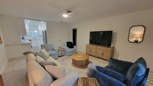 Ein Wohnzimmer mit einer Couch, Sesseln, einem Couchtisch, einem Fernseher und einer Küche im Hintergrund.