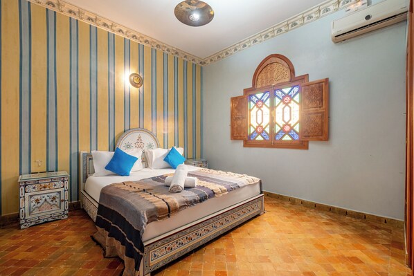 Une chambre à coucher avec un grand lit, une table de chevet, un climatiseur fixé au mur et une fenêtre décorative.