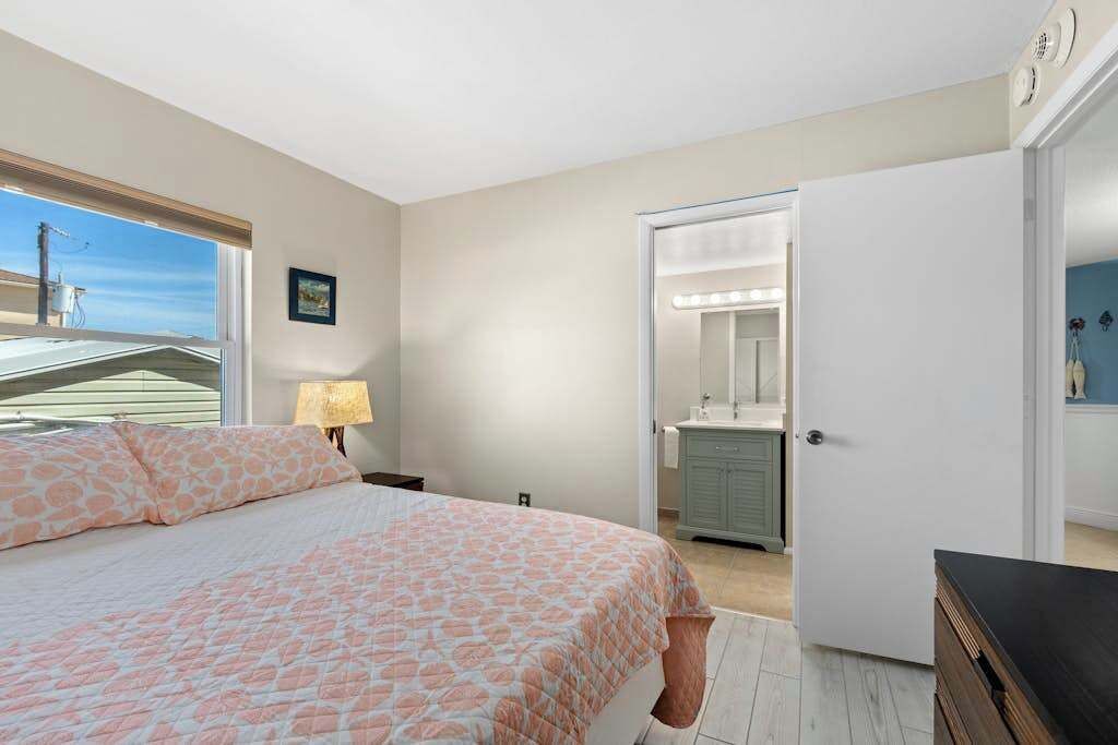 A bedroom with a bed, a nightstand, a lamp, a bathroom with a mirror, and a door leading to another room.