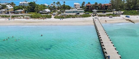 A coastal hotel with a pier extending into turquoise waters, beach access, and a backdrop of residential buildings.