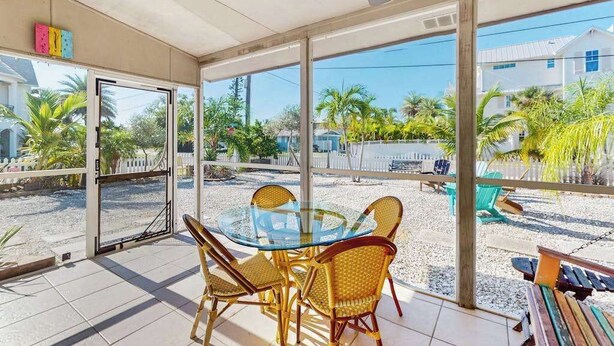 Een overdekt terras met een glazen tafel en rieten stoelen, uitkijkend op een grindgebied met palmbomen en een witte omheining.