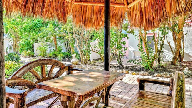 Un espacio de estar al aire libre con techo de paja, mobiliario de madera y jardín de fondo.