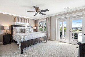 Quarto com cama, ventoinha no teto, mesa de cabeceira, abajur e vista através de uma porta.