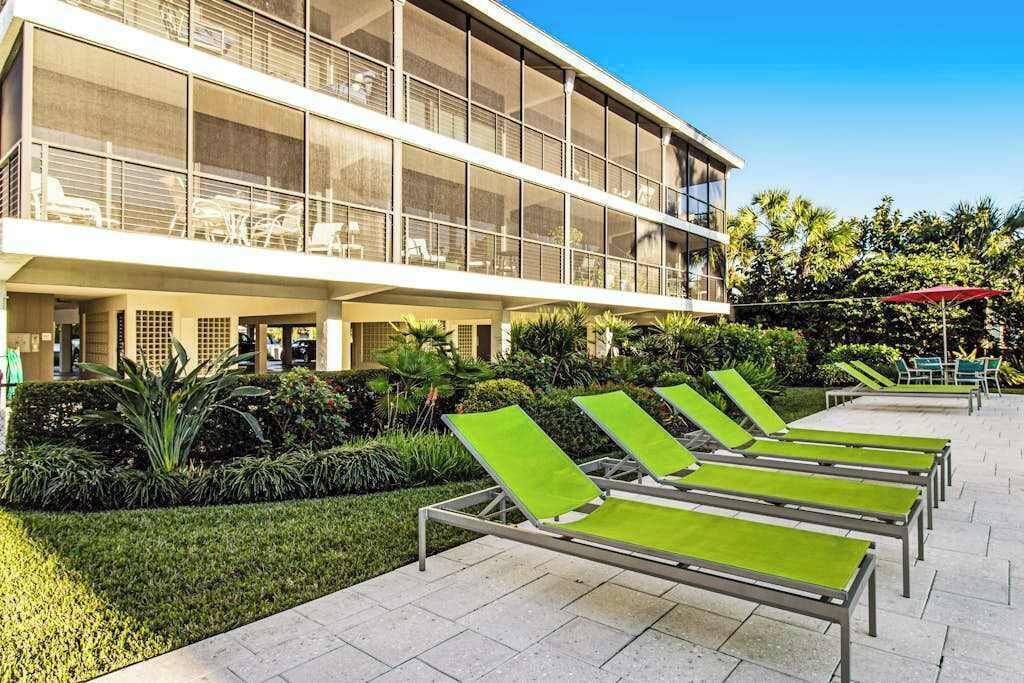 Un edificio moderno con fachada de vidrio, zona de estar al aire libre con sillas verdes y un jardín bien cuidado.