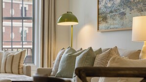 Ein Zimmer mit einer Couch, einem Tisch und einer Lampe. Ein Fenster mit Vorhängen und ein Gemälde an der Wand.