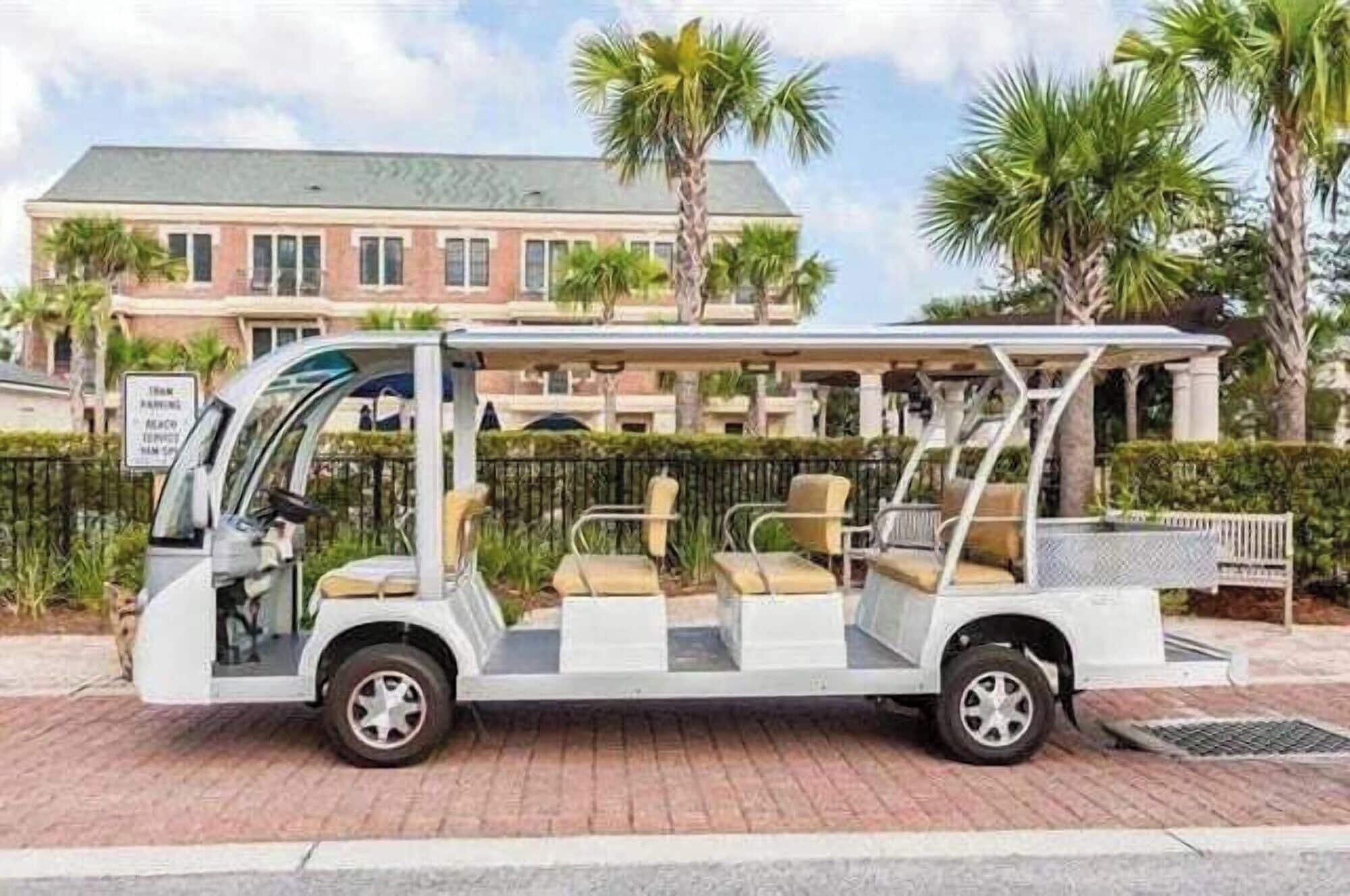 Carro de golf blanco con zona de asientos cubierta y banca, estacionado sobre superficie pavimentada, con un edificio y palmeras al fondo.