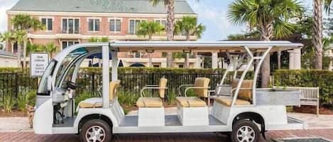 Ein weißer Golfwagen mit überdachter Sitzgelegenheit und einer Bank, geparkt auf einer befestigten Fläche mit einem Gebäude und Palmen im Hintergrund.