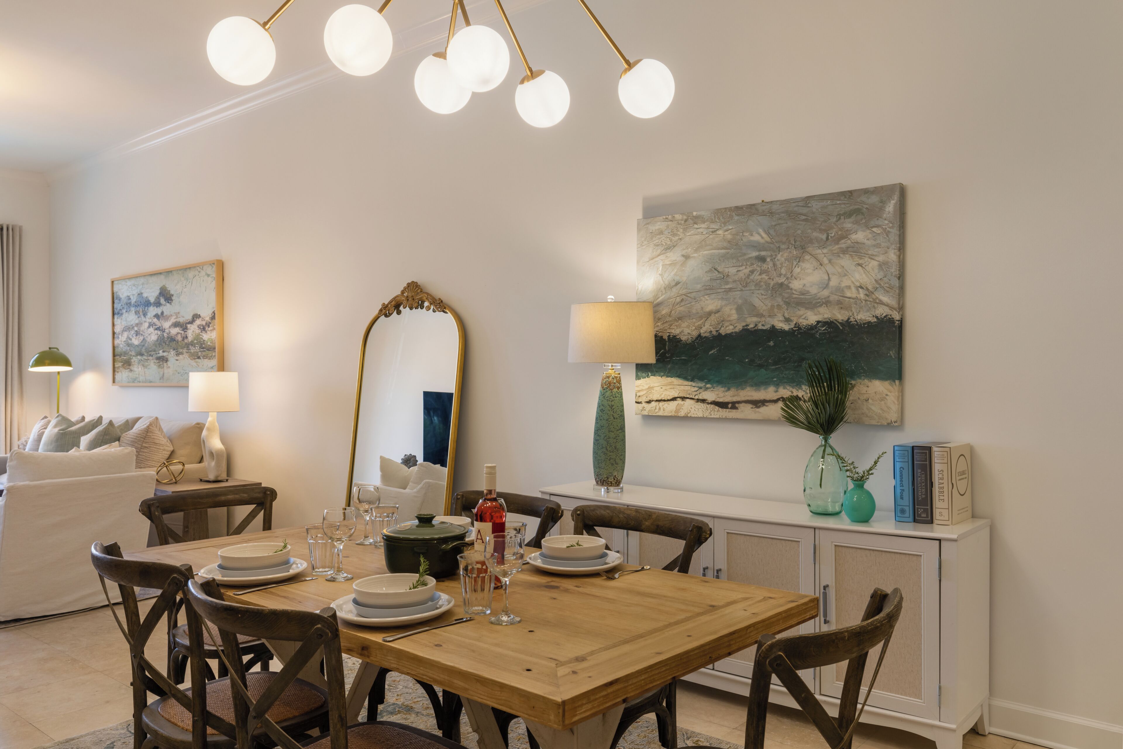A dining area with a wooden table set for four, surrounded by chairs. A large mirror and a painting are on the wall.