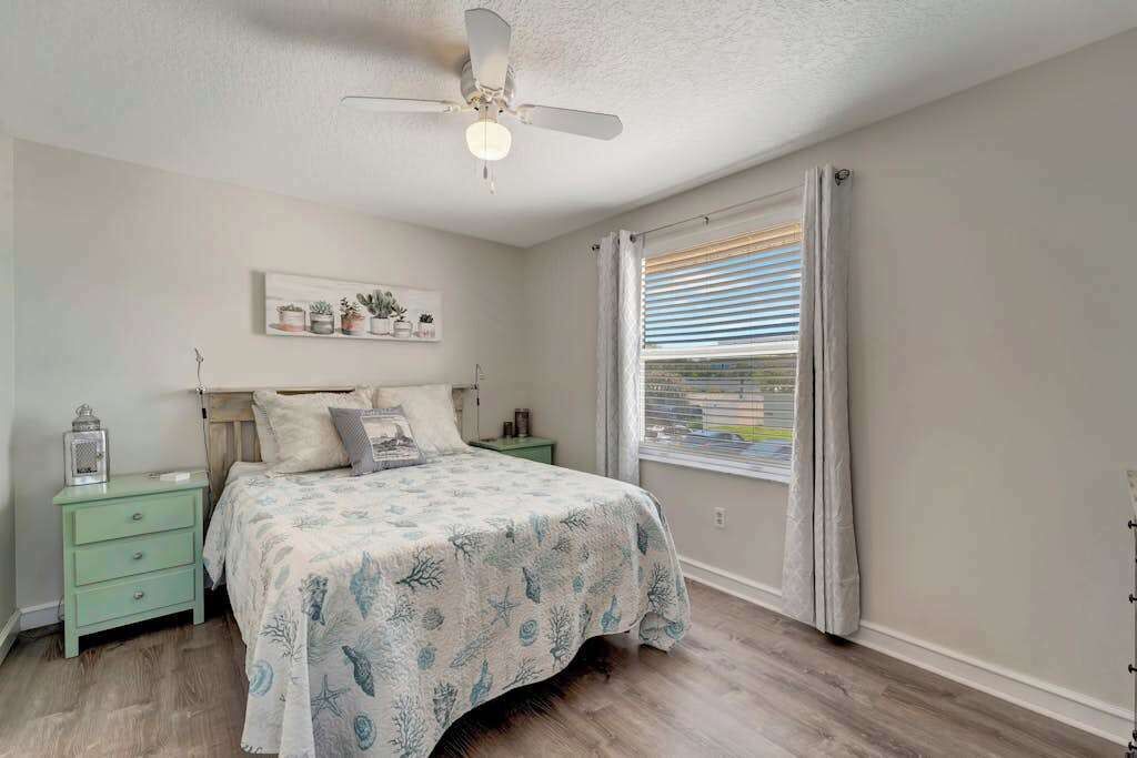 Un dormitorio con cama, mesita de noche, ventilador de techo, ventana con persianas y estante empotrado con objetos decorativos.
