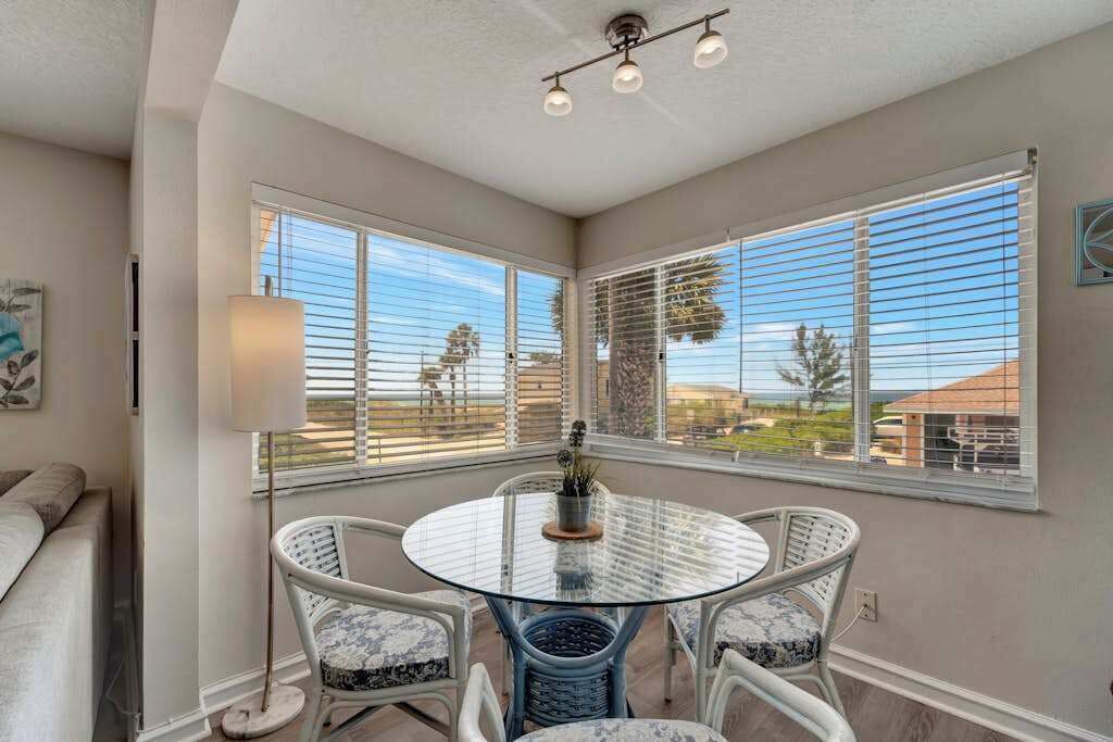 A room with a round glass table, white wicker chairs, a lamp, and large windows with blinds.