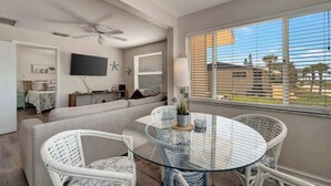 Een moderne woonkamer met een glazen tafel, witte rieten stoelen, een plafondventilator en een aan de muur bevestigde televisie.