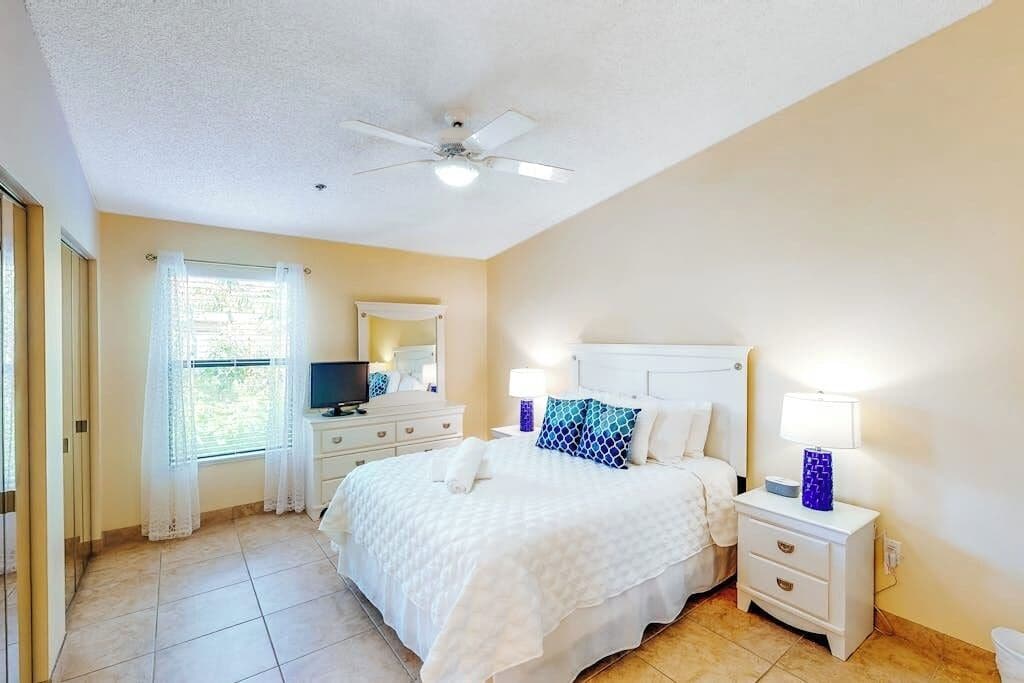 A hotel room with a bed, a television, a ceiling fan, and a window with curtains.