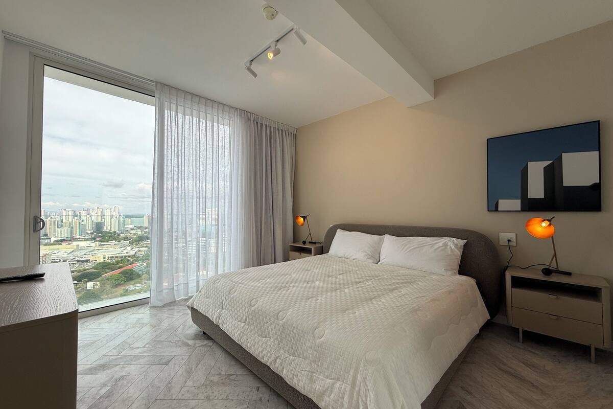 A modern hotel room with a large bed, a city view through floor-to-ceiling windows, a flat-screen TV, and a minimalist decor.