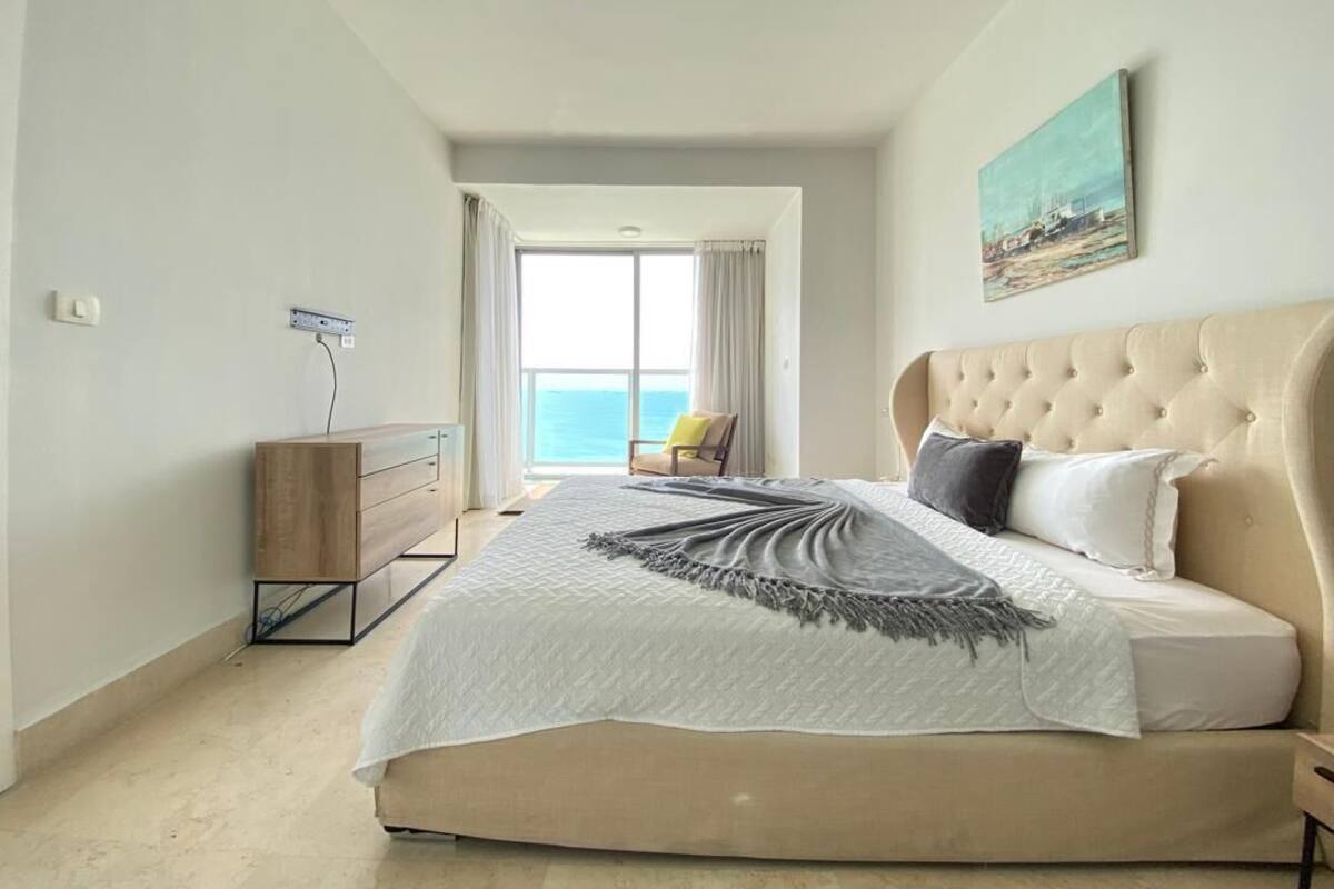 A bedroom with a large bed, a wooden dresser, a chair, and a view of the sea.