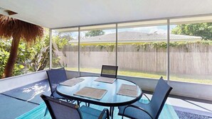 A covered patio with a round table and chairs, large windows, and a view of a fence and greenery. - Charming home w large yard, near Beaches, Sarasota, Bradenton, & the airport (Sarasota)