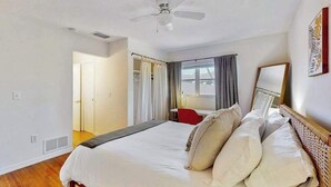 Une chambre d’hôtel avec un lit, un ventilateur de plafond, une fenêtre avec des rideaux, une chaise rouge et un miroir au mur.