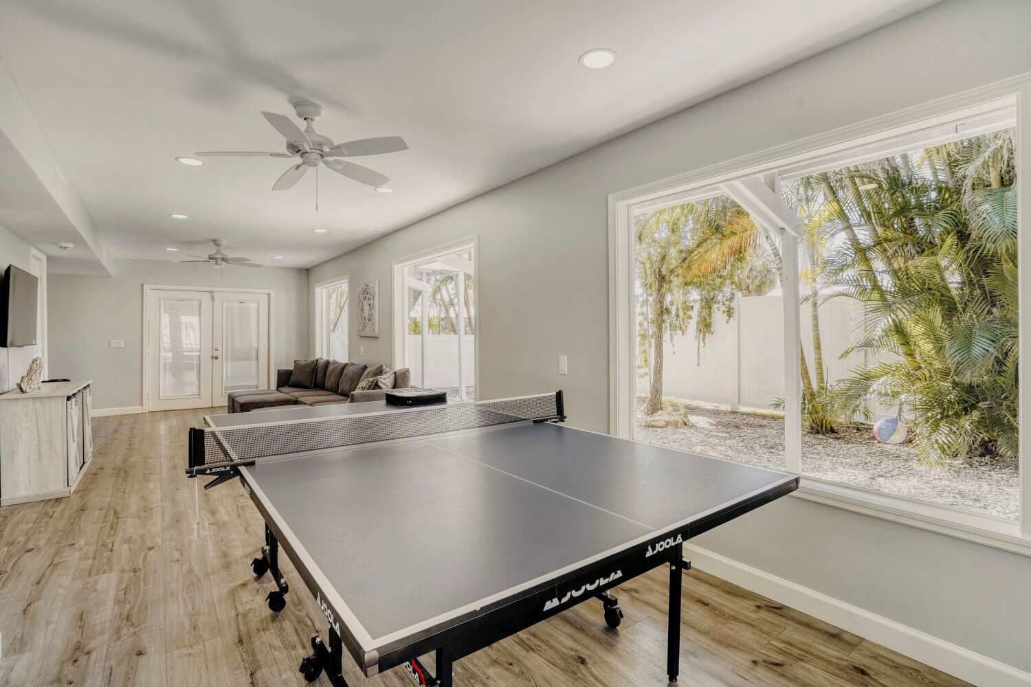 A room with a ping pong table, a sofa, a ceiling fan, and large windows with a view of palm trees.