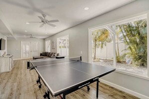 A room with a ping pong table, a sofa, a ceiling fan, and large windows with a view of palm trees. - Relaxing Home with Private Pool, High-Speed WiFi, Central AC, & Washer-Dryer (Anna Maria)