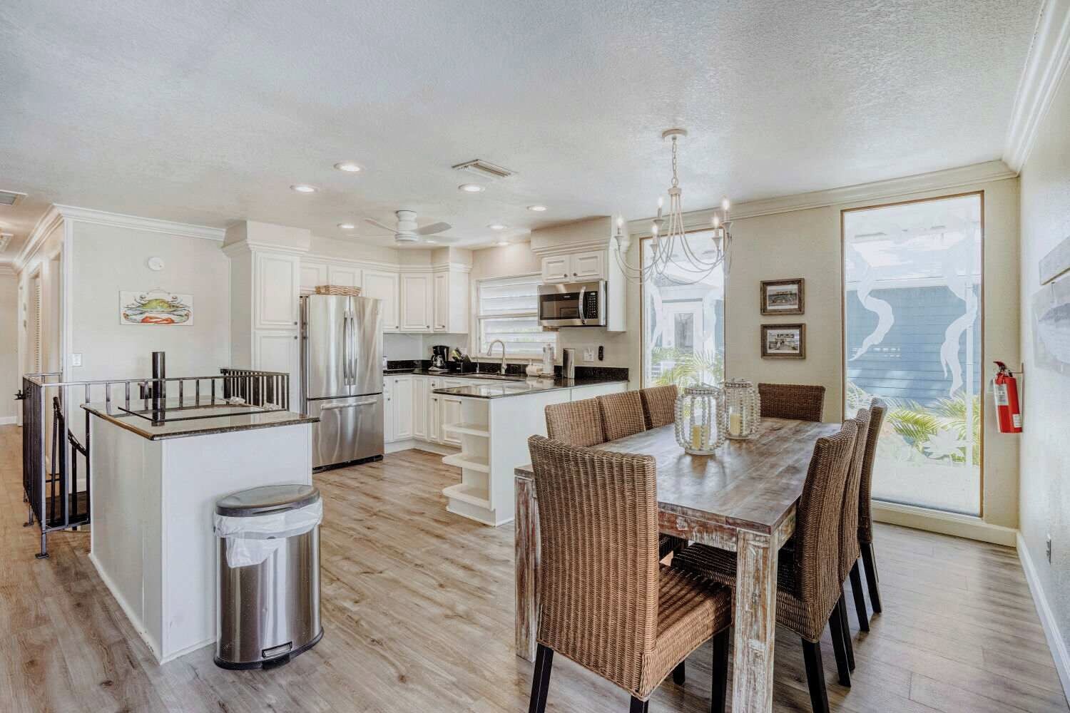 A spacious kitchen with a central island, dining area with wicker chairs, and large windows offering a view.