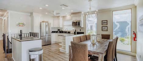 A spacious kitchen with a central island, dining area with wicker chairs, and large windows offering a view.