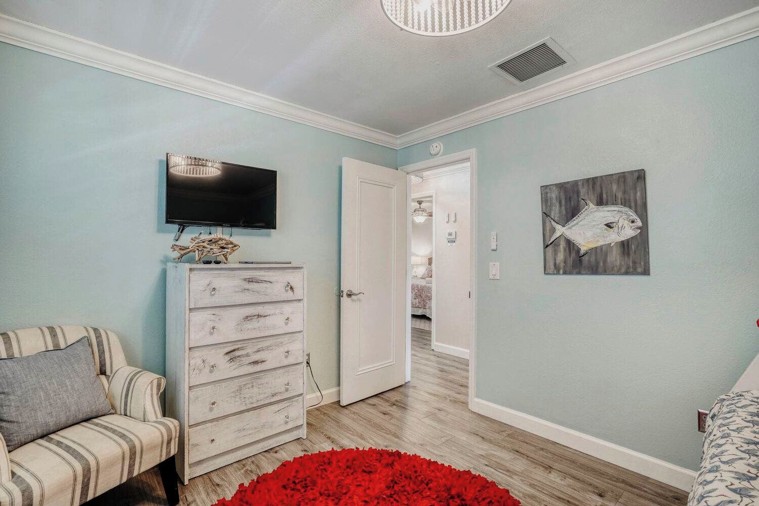 A room with a television mounted above a white dresser, a red rug on the floor, and a fish painting on the wall.