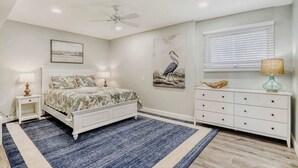 Une chambre à coucher comprenant un lit, des tables de chevet, une commode, un ventilateur de plafond, une fenêtre avec des stores et une œuvre d’art accrochée au mur.