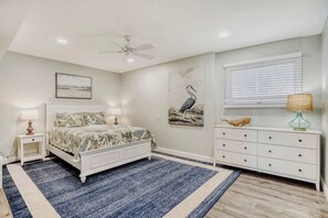 Ein Schlafzimmer mit einem Bett, Nachttischen, einer Kommode, einem Deckenventilator, einem Fenster mit Jalousien und einem Wandbild.