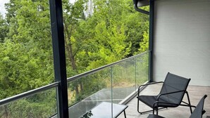 A balcony with two black chairs, a small table, and a glass railing overlooking a forested area.