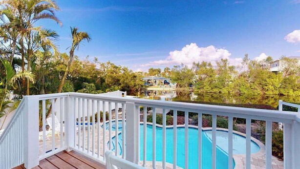 Piscina com deck de madeira e guarda-corpo branco, rodeada por vegetação e céu claro.