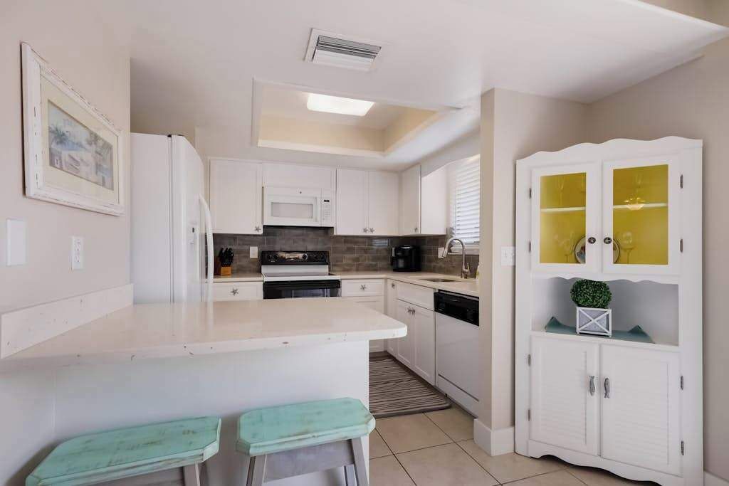 Una cocina moderna con armarios blancos, un microondas, una estufa y un lavamanos. Hay una pequeña isla con taburetes y un mueble con puertas de vidrio.