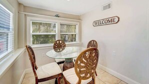 Un comedor con una mesa redonda de vidrio, cuatro sillas de madera con asientos acolchados, una ventana con persianas y un letrero que dice 'WELCOME' en la pared.