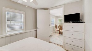 Un dormitorio con cama, televisión sobre una cómoda, ventilador de techo y puerta que da a un balcón.