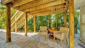 Una pérgola de madera con una escalera, dos sillas de mimbre y una mesita.