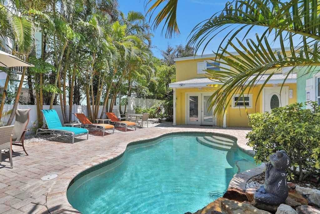 A swimming pool with lounge chairs, palm trees, and a yellow building.