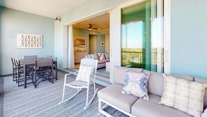 Une pièce comprenant une table à manger, des chaises, un canapé et un fauteuil à bascule. On peut admirer, depuis cette pièce, un balcon avec une balustrade.