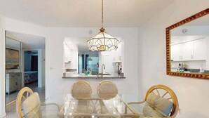 Zona pranzo con tavolo in vetro e sedie in vimini, un lampadario e una cucina visibile attraverso una porta aperta.