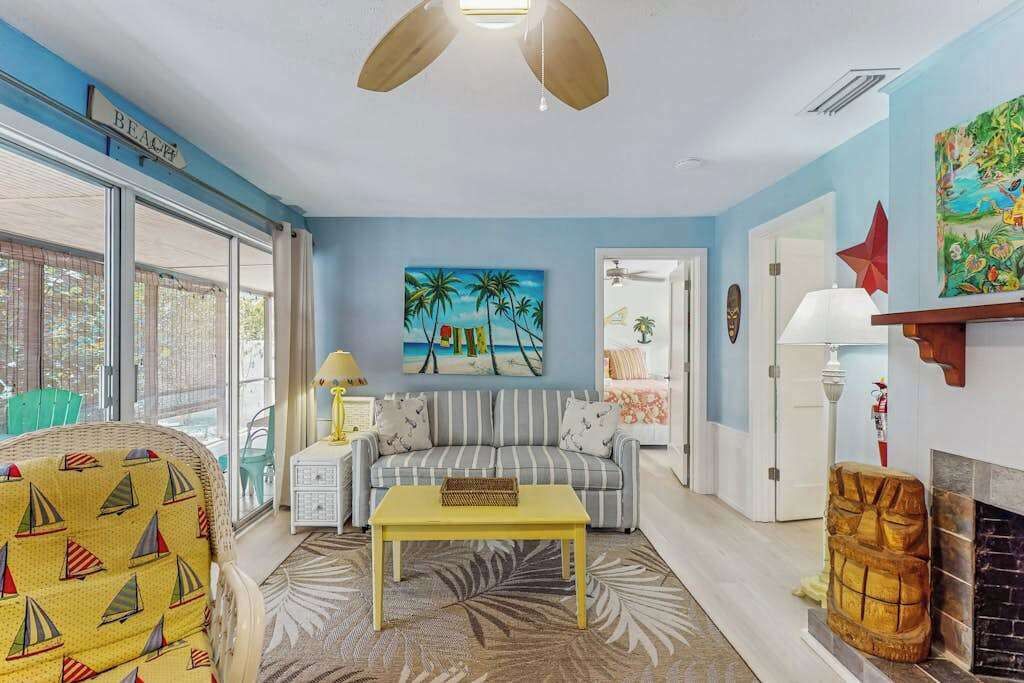A living room with a sofa, a yellow coffee table, a patterned armchair, a side table with a lamp, and a painting of palm trees.