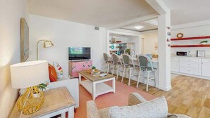 Una sala de estar con sofá, mesa de centro y televisor. Se observa un comedor con sillas y una cocina al fondo.