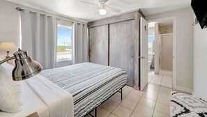 Une chambre à coucher avec un lit, un ventilateur de plafond, une lampe, une vue sur l’extérieur et une porte donnant sur la salle de bain.