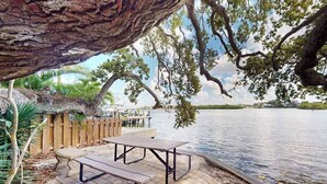 Ein Picknicktisch unter einer großen Baum, am See gelegen, mit Holzzaun und Stühlen im Hintergrund.
