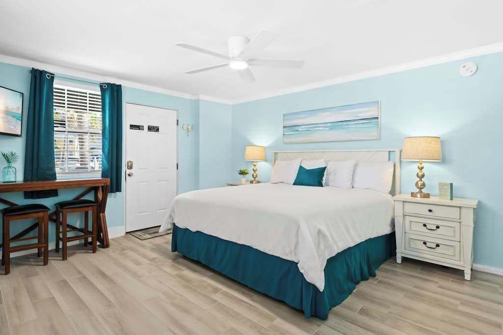 A bedroom with a bed, two bedside tables, a desk with chairs, a ceiling fan, and a window with curtains.