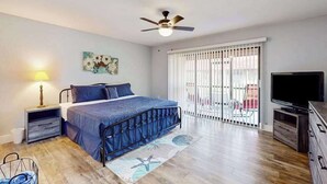 Quarto com cama, ventoinha no teto, TV, janela com persianas e varanda.