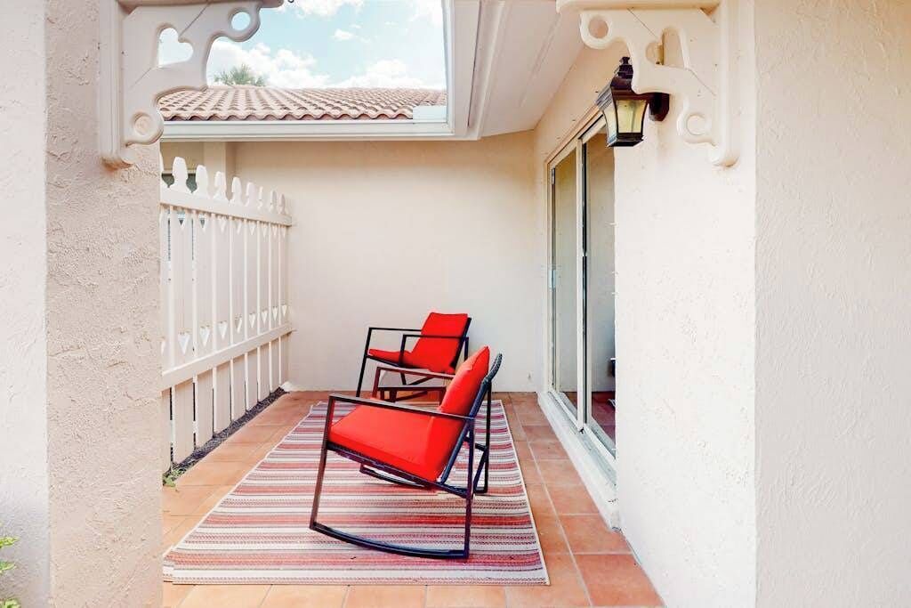 A balcony with two red chairs, a striped rug, a white fence, and a view of a building with a tiled roof.