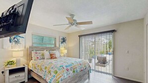 Ein Schlafzimmer mit Bett, Nachttisch, Deckenventilator, Fenster mit Jalousien und Blick ins Freie.